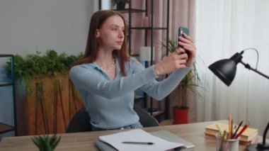 Young woman blogger taking selfie portraits on smartphone for social media vlog while sitting at table working from home office. Girl businesswoman freelancer study, e-learning, remote job online