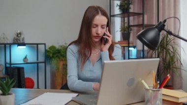 Young woman freelancer sits at desk at home office answering to client on mobile phone call, using laptop computer. Conversation. Adult girl student study working online remote distance job, talking