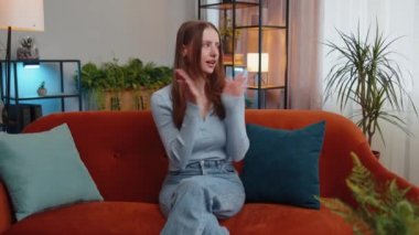 Happy young woman shouting, celebrating success, winning, goal achievement, good news, raising fists in gesture I did it, lottery luck at home apartment. Joyful girl indoors in living room on couch