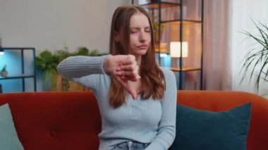 Dislike. Upset young woman showing thumbs down sign gesture, expressing discontent, disapproval, dissatisfied bad work at modern home apartment indoors. Displeased girl in living room sitting on sofa