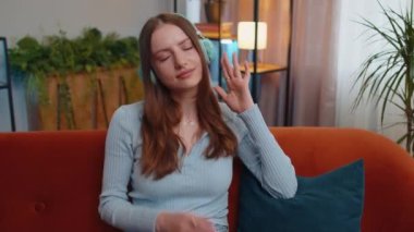 Portrait of happy relaxed overjoyed young woman in wireless headphones dancing on couch at home choosing listening favorite energetic disco music in smartphone entertaining. People weekend activities