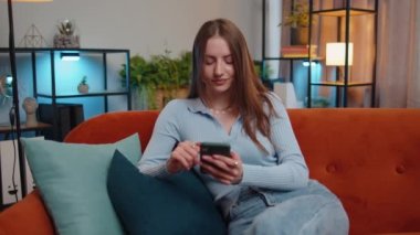 Kanepede oturan genç bir kadın modern bir evde cep telefonu tebessümü kullanıyor. Güzel esmer bir kız akıllı telefon sosyal medya uygulamalarında mesajlarını paylaşıyor, rahatlama filmi izliyor.