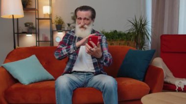 Senior old grandfather man wearing earphones making phone online conversation call with family sitting at home living room couch. Elderly retired grandpa pensioner enjoying mobile loudspeaker talking