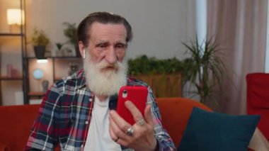 Yaşlı büyükbaba kulaklık takıyor. Evde oturma odasındaki kanepede oturan aileyle telefonda sohbet ediyor. Emekli yaşlı büyükbaba emeklisi. Mobil hoparlörden keyif alıyor.
