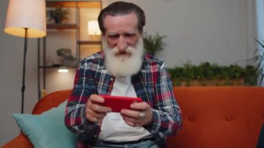 Worried senior old grandfather man enthusiastically playing racing shooter online video games on mobile phone. Elderly grandpa using smartphone gadget app with drive simulator at home sitting on couch
