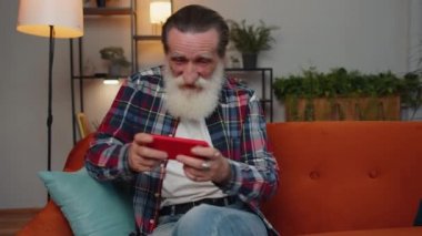 Worried senior old grandfather man enthusiastically playing racing shooter online video games on mobile phone. Elderly grandpa using smartphone gadget app with drive simulator at home sitting on couch