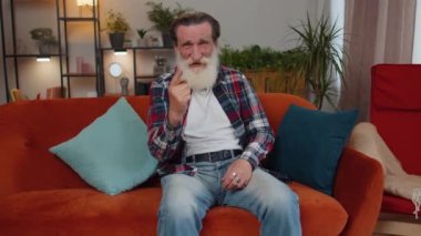 Heyecanlı yaşlı büyükbaba bağırıyor, başarıyı kutluyor, kazanıyor, gol başarısı, iyi haber, el hareketleriyle yumruk kaldırıyor. Olgun adam kanepede odada.