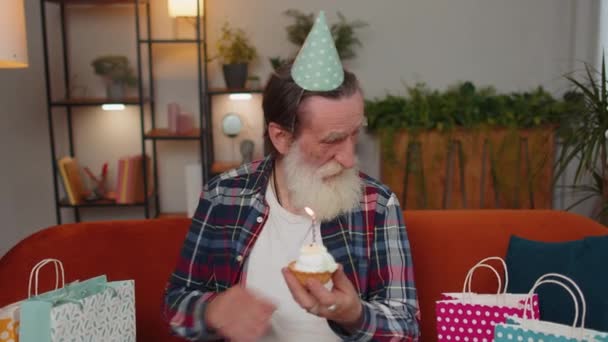 할아버지가 생일을 축하하는 모자를 케이크 위에서 촛불을 불어서 축하하고 집에서 — 비디오