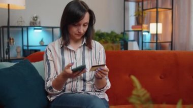 Young woman using credit bank card and smartphone while transferring money, purchases online shopping, order food delivery at modern home apartment indoors. Pretty girl in living room sitting on couch