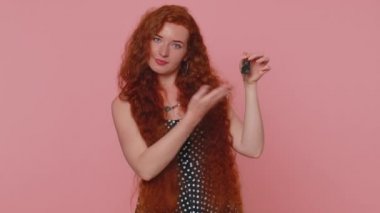 Redhead young woman real estate agent lifting hand showing the keys of new home house apartment, buying or renting property, mortgage loan. Red hair ginger girl isolated on pink studio wall background