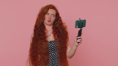 Wow reaction. Impressed redhead woman blogger taking selfie on mobile phone selfie stick, communicating video call online with subscribers. Young girl ginger isolated alone on pink studio background