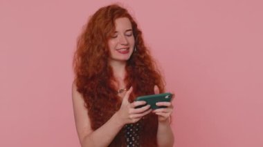 Endişeli komik bağımlı kızıl kadın cep telefonuyla yarış oyunu oynuyor. Genç kızıl saçlı kız pembe stüdyo arka planında izole edilmiş sürücü simülatörlü akıllı telefon aygıtı kullanıyor.