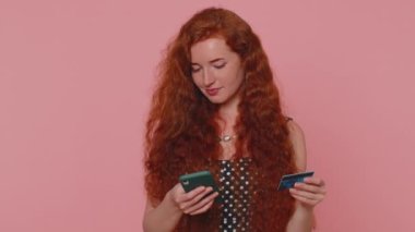 Sincere redhead woman customer using credit bank card and smartphone while transferring money, purchases online shopping, payment. Internet finance. Young girl isolated alone on pink wall background