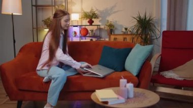 Young redhead teenager child girl freelancer enters the home room and sits down on couch, opens laptop. Ginger children kid works on notebook, sends messages, makes online purchases, watching movies