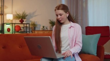 Young teen child girl sitting on couch, looking at laptop, making video webcam conference call with friends or family, enjoying pleasant conversation. Ginger kid laughing, waving hello alone in home