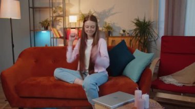Excited young teen child girl shouting, celebrating success, winning, goal achievement, good news, raising fists in gesture I did it, lottery luck at home room. Redhead kid indoors in room on couch