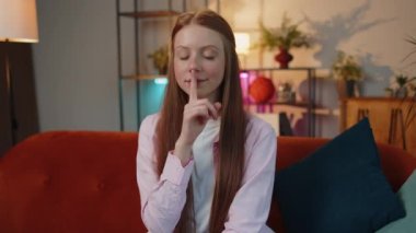 Shh be quiet please. Young redhead child girl presses index finger to lips makes silence hush gesture sign do not tells gossip secret. Teen freckles kid at home apartment living room sitting on couch