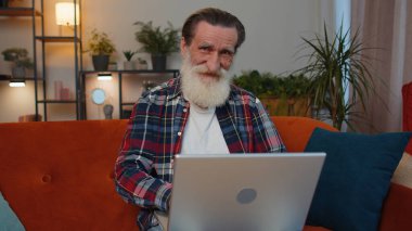 Ev deki koltukta dizüstü bilgisayar çığlığıyla aşırı mutlu yaşlı büyükbaba zaferi kutlarken ellerini kaldır piyangodan para kazan. Heyecanlı yaşlı adam internette iyi haberler alıyor.