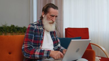 Yaşlı büyükbaba, sınıftaki kanepede dizüstü bilgisayarla çalışıyor. Yaşlı emekli büyükbaba defter üzerinde çalışıyor, mesajlar gönderiyor, online siparişler veya yemek siparişleri veriyor, film seyrediyor.