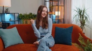 Mutlu genç bir kadın bağırıyor, başarıyı kutluyor, kazanıyor, gol başarısı, iyi haber, iyi haber, el hareketleriyle yumruk kaldırıyor. Ben yaptım, evde piyango şansı. Oturma odasında kanepede neşeli bir kız.