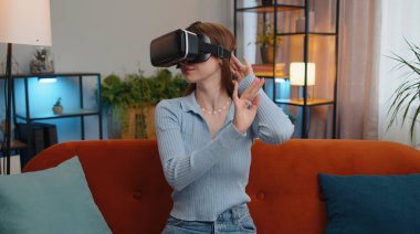 Genç bir kadın sanal gerçeklik teknolojisi VR uygulama kulaklığı kullanarak 3D 360 video oyunu simülasyonu yapıyor ve modern ev dairesinde film izliyor. Gözlüklü güzel kız kanepede oturuyor.
