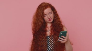 Akıllı telefon görünümlü mutlu kızıl saçlı genç kadın içten bir sevinç gösterisi kazanıyor, para dolar banknotları alıyor, başarı piyangosunda şans getiriyor. Kapalı alan stüdyosundaki kızıl kız pembe duvar arkasında izole edilmiş.