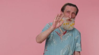 Handsome senior man in t-shirt smiling friendly at camera and waving hands gesturing hello or goodbye, welcoming with hospitable expression. Happy grandfather indoors isolated on pink wall background