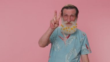 Senior man with flowers in gray-haired beard showing victory sign, hoping for success and win, doing peace gesture, smiling with kind optimistic expression. Elderly grandfather on pink wall background