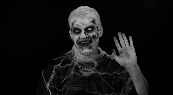 Creepy man with bloody scars face, Halloween zombie make-up. Scary wounded undead guy smiling friendly at camera and waving hands gesturing hello or goodbye, welcoming with hospitable expression