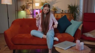 Happy relaxed overjoyed child girl in wireless headphones dancing on couch at home choosing listening favorite energetic disco rock n roll music in smartphone entertaining. People weekend activities