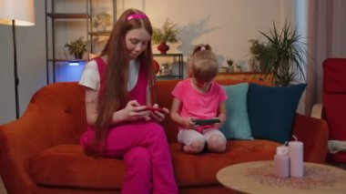 Worried funny girls friends siblings enthusiastically playing shooting video games on smartphone. Happy two female child kids family on couch at home, using smartphone gadget app with drive simulator