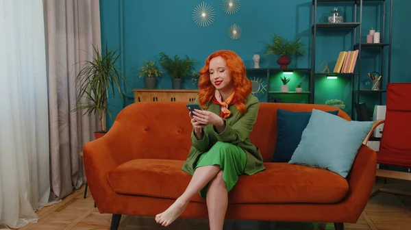 Cheerful woman sitting on sofa uses mobile phone smile at modern home apartment. Happy redhead girl texting share messages content on smartphone social media applications online, watching relax movie