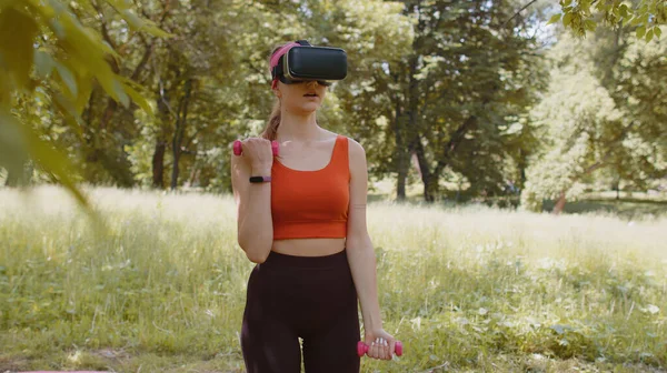 Athletic girl in VR headset helmet making fitness exercises with dumbbells outdoors in sunny park. Online workout. Young woman in sportswear watching virtual reality 3D video sport bodybuilder game