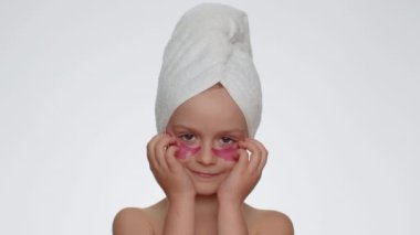 Beautiful young smiling child girl in towel on head applying pink cosmetic patches under eyes. Teenager kid face skin care treatment. Female portrait on white background. Perfect natural fresh clean
