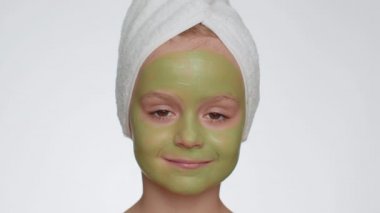 Smiling child girl in towel on head applying cleansing moisturizing green cucumber vitamin facial mask. Teenager kid face skin care treatment, natural cosmetics. Female portrait. Perfect fresh clean