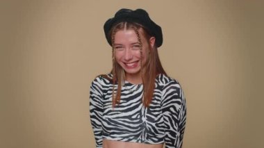 Smiling playful happy woman blinking eye, looking at camera with toothy smile, winking and flirting, expressing optimism. Adult stylish female girl isolated alone on beige studio background indoors