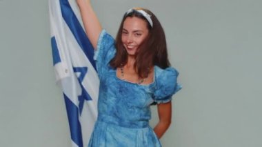 Lovely pretty young woman in blue dress waving, wrapping in Israel national flag, celebrating Independence day. Patriotism. Adult stylish female girl isolated alone on gray studio background indoors