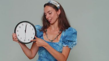 It is your time. Pretty young woman in blue princess dress showing time on wall office clock, ok, thumb up, approve, pointing finger at camera. Stylish female girl on gray studio background indoor