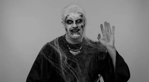 Creepy man with bloody scars face, Halloween zombie make-up. Scary wounded undead guy smiling friendly at camera and waving hands gesturing hello or goodbye, welcoming with hospitable expression