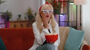 Excited senior grandmother sitting on sofa eating popcorn and watching interesting tv serial, sport game film, online social media movie content online at home. Woman enjoying domestic entertainment