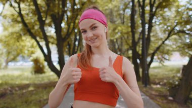 Atletik formda, gülümseyen koşucu kız eğitiminin portresi kameraya bakıyor, maraton başarısından mutlu. Parkta egzersiz kardiyolojisi. Kadın başparmağını kaldırır bir şeye katılır ya da olumlu cevap verir