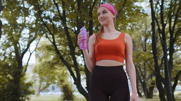 Athletic fitness sport runner girl drinking water from bottle after training exercising. Workout cardio in park at morning. Young woman jogger enjoy healthy lifestyle. Active sportswoman activities
