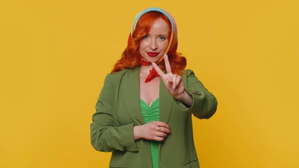 Redhead young woman in green jacket showing victory sign, hoping for success and win, doing peace gesture, smiling with kind optimistic expression. Ginger girl indoors isolated on yellow background