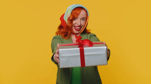 Smiling redhead young woman presenting birthday gift box stretches out hands, offer wrapped present career bonus, celebrating party. Red hair ginger girl isolated alone on yellow studio background