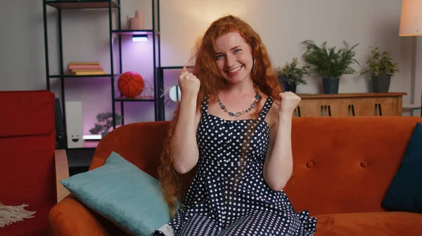 Redhead woman with freckles shouting, celebrating success, winning, goal achievement, good news, raising fists in gesture I did it, lottery luck at home. Ginger girl indoors in living room on couch
