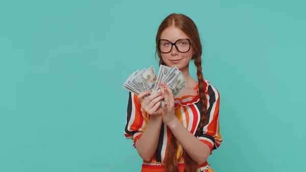 Redhead young girl holding fan of cash money dollar banknotes celebrate dance, success business career, lottery game winner, big income, wealth. Red hair teenager child alone on blue studio background