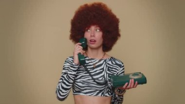 Hey you, call me back. Pretty young woman with brown lush wig talking on wired vintage telephone of 80s, says hey you call me back. Adult stylish female girl isolated on beige studio background indoor