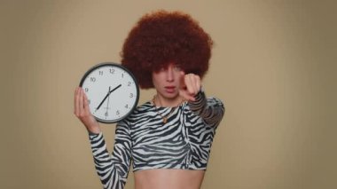 It is your time. Pretty young woman with brown lush wig showing time on wall office clock, ok, thumb up, approve, pointing finger at camera. Adult stylish female girl on beige studio background indoor