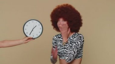 Young woman in brown lush wig with anxiety checking time on clock, running late to work, being in delay, deadline. Adult stylish female girl looking at hour, minutes, worrying to be punctual, indoor