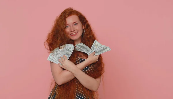 Redhead young woman holding fan of cash money dollar banknotes celebrate dance, success business career, lottery game winner, big income wealth. Red hair ginger freckles girl on pink studio background